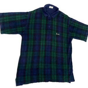 May include: A short-sleeved polo shirt featuring a dark blue and green plaid pattern. The shirt has a solid dark blue collar, a three-button placket, and the word Harrods embroidered in white thread on the left chest area. A white brand label is visible inside the collar.
