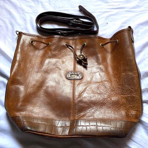 May include: A brown leather bucket bag with a shoulder strap. The bag has a drawstring closure and a textured bottom panel. The leather has a worn appearance. The strap is a dark brown color.