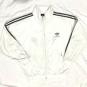 May include: White track jacket with black stripes on the sleeves and shoulders. The jacket has a full zip and the Adidas logo on the chest. The jacket is made of a shiny material.