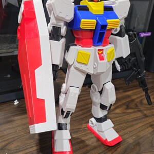 May include: A detailed, multi-colored Gundam model, primarily white with red, blue, yellow, and black accents. The model is holding a red and white shield and a black weapon. The figure is standing on a wooden surface.