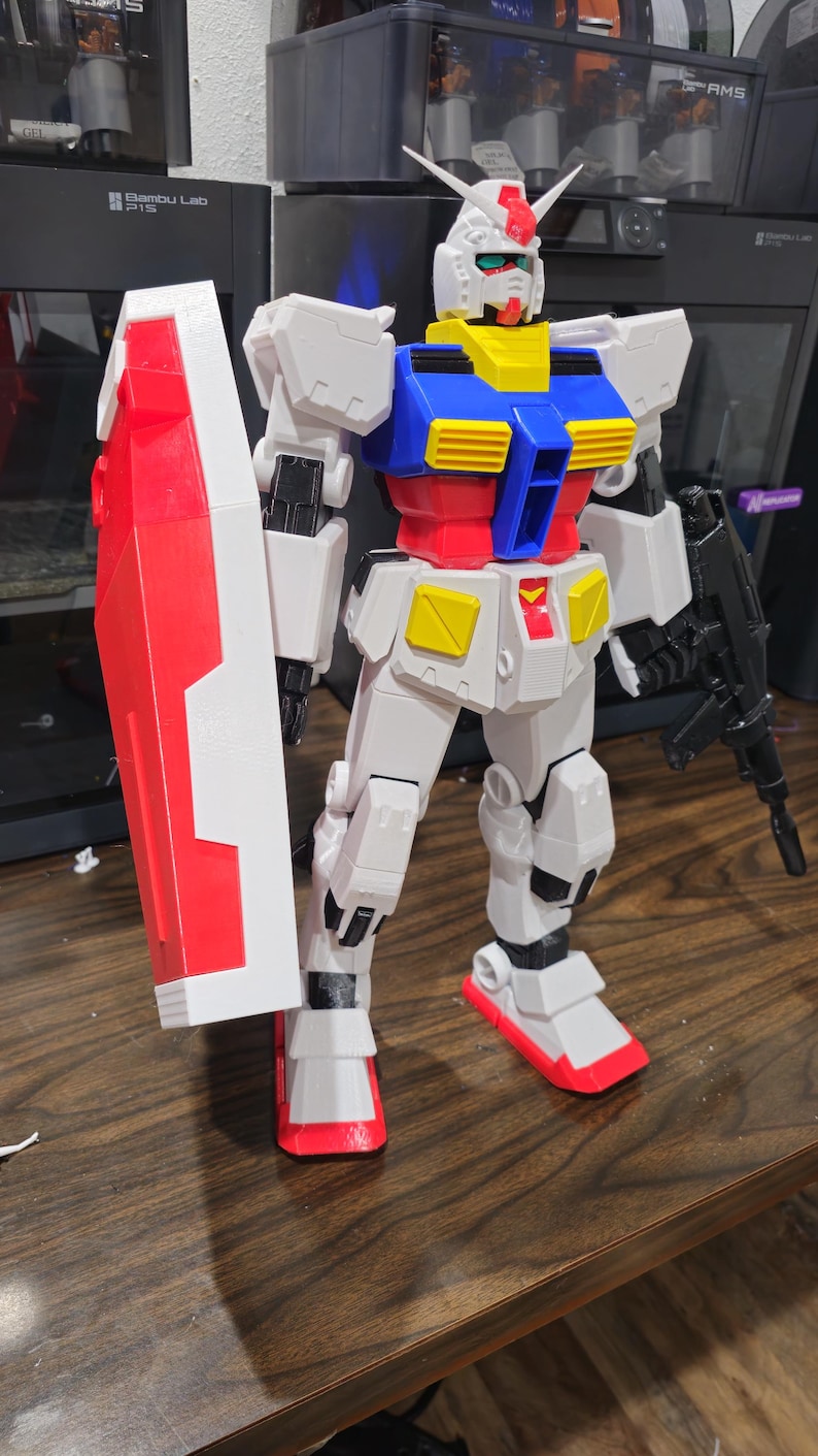 May include: A detailed, multi-colored Gundam model, primarily white with red, blue, yellow, and black accents. The model is holding a red and white shield and a black weapon. The figure is standing on a wooden surface.