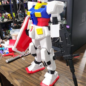 May include: A white, red, blue, and yellow Gundam model stands on a wooden surface. The model is holding a black rifle and a red and white shield. The model is a detailed representation of a robot.