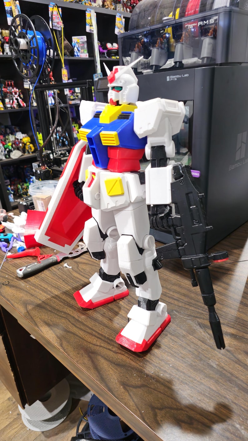 May include: A white, red, blue, and yellow Gundam model stands on a wooden surface. The model is holding a black rifle and a red and white shield. The model is a detailed representation of a robot.