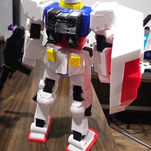 May include: A white, blue, and red robot action figure with a shield and a gun. The robot has a white head with two antennae, a yellow collar, and a black chest. The robot is standing on a wooden surface.