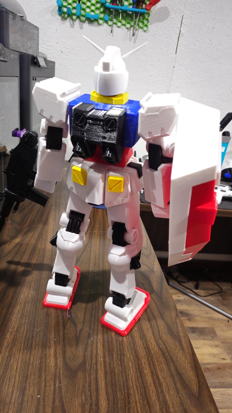 May include: A white, blue, and red robot action figure with a shield and a gun. The robot has a white head with two antennae, a yellow collar, and a black chest. The robot is standing on a wooden surface.