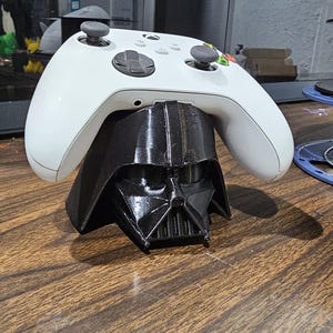 May include: A white Xbox controller rests on a black Darth Vader helmet-shaped stand. The controller has black buttons and gray joysticks. The stand is on a wooden surface.