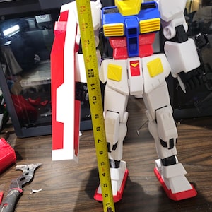 May include: A large, multi-colored Gundam model, approximately 19 inches tall, with a red, white, blue, and yellow color scheme. The model is standing upright with a shield. The image is taken indoors.