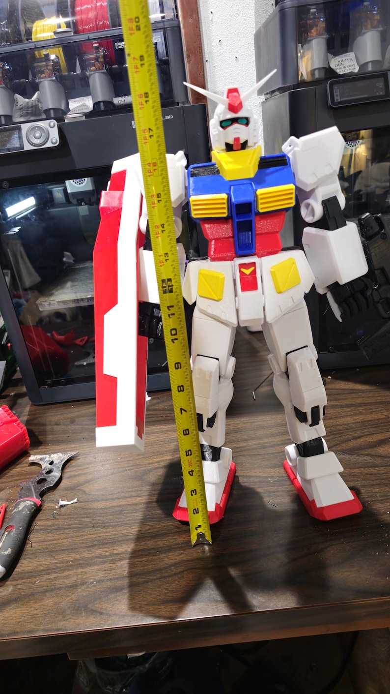 May include: A large, multi-colored Gundam model, approximately 19 inches tall, with a red, white, blue, and yellow color scheme. The model is standing upright with a shield. The image is taken indoors.
