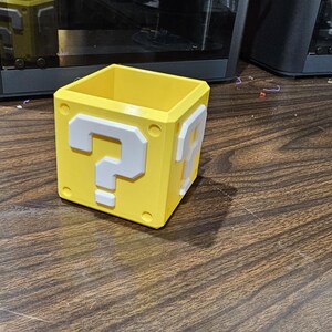 May include: A yellow, cube-shaped container with white question marks on two sides. The container is a 3D-printed item, likely for desk organisation or storage. The design is inspired by a video game.