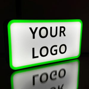 Puede incluir: Un letrero rectangular con una superficie blanca y las palabras "YOUR LOGO" en letras negras en negrita. El letrero tiene un borde verde brillante y se refleja en una superficie negra brillante.