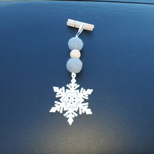 May include: A white snowflake ornament with gray felt balls and wooden beads hanging from a wooden clothespin.
