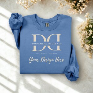 May include: A light blue sweatshirt with the text "Dopamine Digitals" and "Your Design Here" in a light beige color. The sweatshirt is folded on a marble surface, with dried flowers in the background.