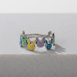 May include: A silver ring featuring a repeating pattern of colorful bunny faces. Each bunny face is a different color, including green, yellow, purple, and blue. The ring is set against a neutral background.