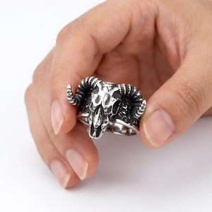 May include: A silver-colored ring featuring a detailed ram skull design. The ring has prominent curved horns and intricate detailing on the skull. The ring is held in a hand against a white background, showcasing the gothic or biker-inspired jewelry.