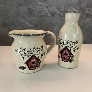 May include: A cream-colored ceramic pitcher and bottle set. Both pieces feature a birdhouse design with red, brown, and green accents. The pitcher has a handle and a ruffled rim with blue trim. The bottle is tall and slender, with a similar design.