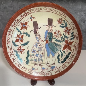May include: A decorative ceramic plate with a hand-painted scene of two figures in period clothing, surrounded by floral and botanical designs. The plate has a brown rim and features text in a script font. The plate is displayed on a wooden stand.