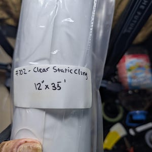 May include: A roll of clear static cling film, sealed in a transparent plastic bag. The label reads "#102 - Clear Static Cling" and indicates dimensions of 12" x 35'. The film appears to be rolled up inside the bag.