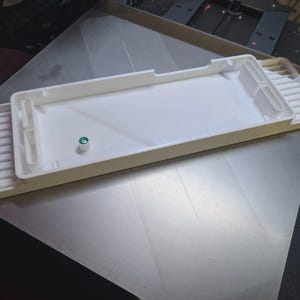 May include: A white plastic keyboard case with a rectangular main section and an attached tray with ridges. A small green and white component is visible. The case is on a silver surface.