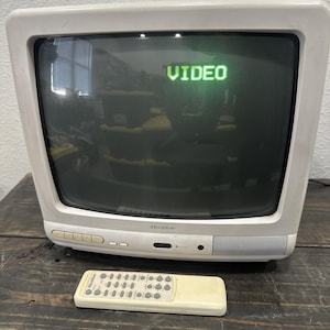 Könnte beinhalten: Cremefarbener Vintage-Fernseher von Quasar mit dem grünen Wort "VIDEO" auf dem Bildschirm. Der Fernseher hat eine passende Fernbedienung. Der Fernseher steht auf einer dunklen Holzoberfläche.