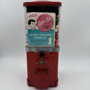 Może przedstawiać: Zabytkowy czerwony automat do gumy do żucia z przezroczystym szklanym wyświetlaczem. Automat posiada etykietę "Rain-Blo Bubble Gum" z postacią z kreskówki. Widoczny jest tekst "Blows Colored Bubbles". Automat wydaje gumy do żucia za 1 cent.