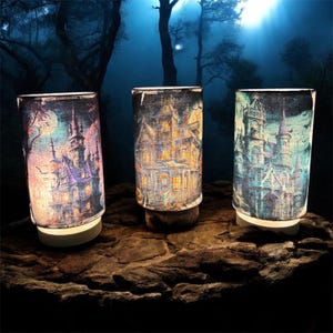 May include: Three cylindrical lamps with illuminated haunted house designs. Each lamp features a different color scheme: purple and pink, yellow and orange, and teal and blue. The lamps are set on a stone surface against a dark, atmospheric background.