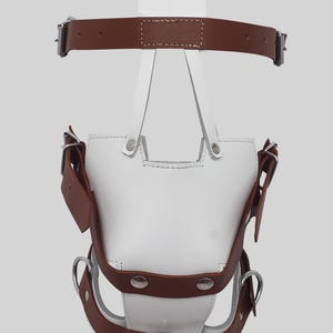 May include: A white leather bag with brown leather straps and silver-tone metal hardware. The bag has a unique design with multiple straps and a handle. The bag is designed to be worn on the body.