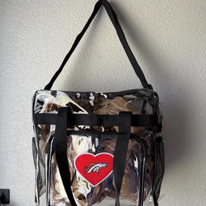 May include: Clear tote bag with black handles and trim. Features a red heart-shaped patch with a white horse logo. Includes side mesh pockets and a camouflage pattern inside the bag.