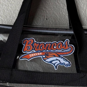 May include: A clear plastic tote bag featuring black nylon straps and a prominent embroidered patch of the Denver Broncos logo. The patch displays the team name in orange and blue script above a stylized white and orange horse head emblem.