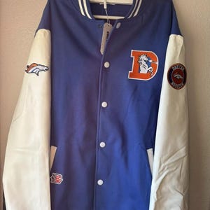 May include: A blue and white varsity jacket with the Denver Broncos logo on the left sleeve and a large "D" with a horse on the right side. The jacket has white sleeves and a white collar with a blue stripe.