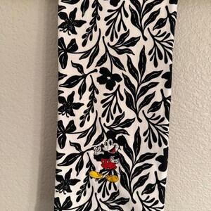Puede incluir: Toalla de cocina blanca con un patr&oacute;n floral negro y un Mickey Mouse bordado en rojo y negro.