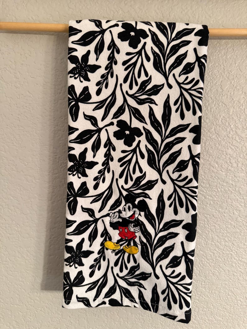 Puede incluir: Toalla de cocina blanca con un patr&oacute;n floral negro y un Mickey Mouse bordado en rojo y negro.