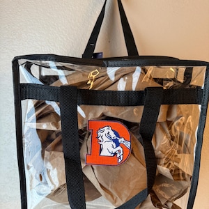 May include: A square, clear plastic tote bag with black fabric trim and black handles. The front features an orange letter D patch with a white horse graphic inside. The bag is filled with crumpled brown paper, visible through the transparent material.