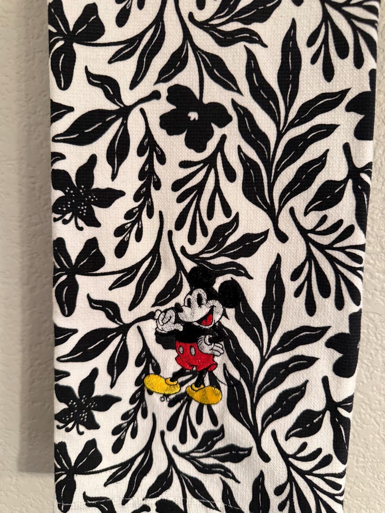 Puede incluir: Toalla de cocina blanca con un patr&oacute;n floral en blanco y negro. Mickey Mouse est&aacute; bordado en el centro de la toalla.