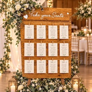 May include: A rustic wooden wedding seating chart board featuring the white text Take your Seats, with twelve white cards clipped to the wood, each listing table numbers and guest names, decorated with white flowers and green eucalyptus leaves.