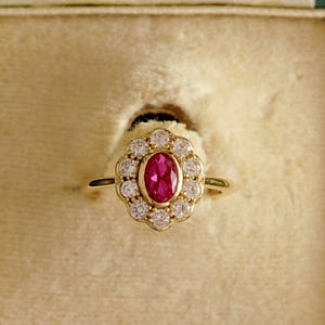 May include: A gold ring featuring an oval ruby centre stone, encircled by a halo of small diamonds. The ring is presented in a vintage green jewellery box with a cream interior. The ring's setting and band are gold.