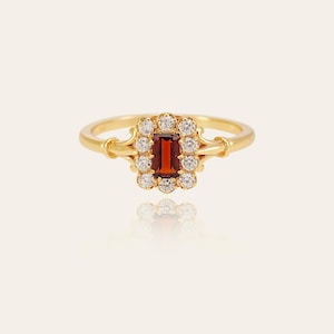 May include: A gold ring with a rectangular, deep red gemstone at its centre, encircled by a halo of small, clear gemstones. The ring's band is polished gold, and the setting has an ornate, vintage-inspired design.