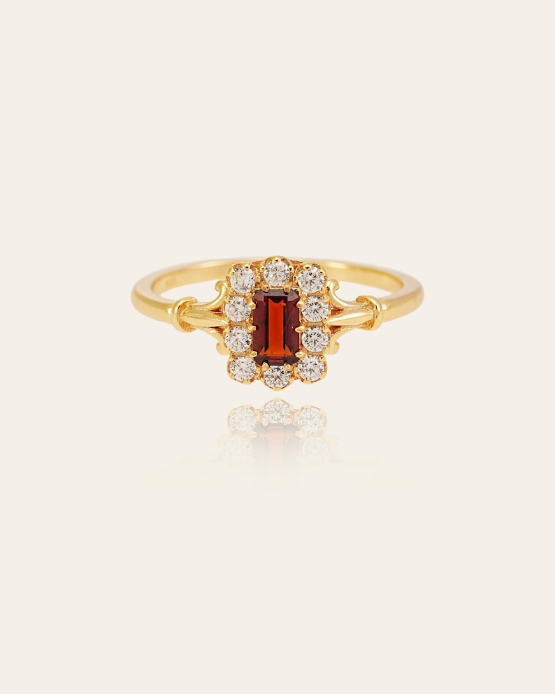 May include: A gold ring with a rectangular, deep red gemstone at its centre, encircled by a halo of small, clear gemstones. The ring's band is polished gold, and the setting has an ornate, vintage-inspired design.
