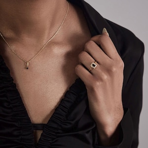 May include: A gold necklace with a small black pendant and a gold ring with a black stone. The ring has a decorative gold setting. The person is wearing a black satin top.