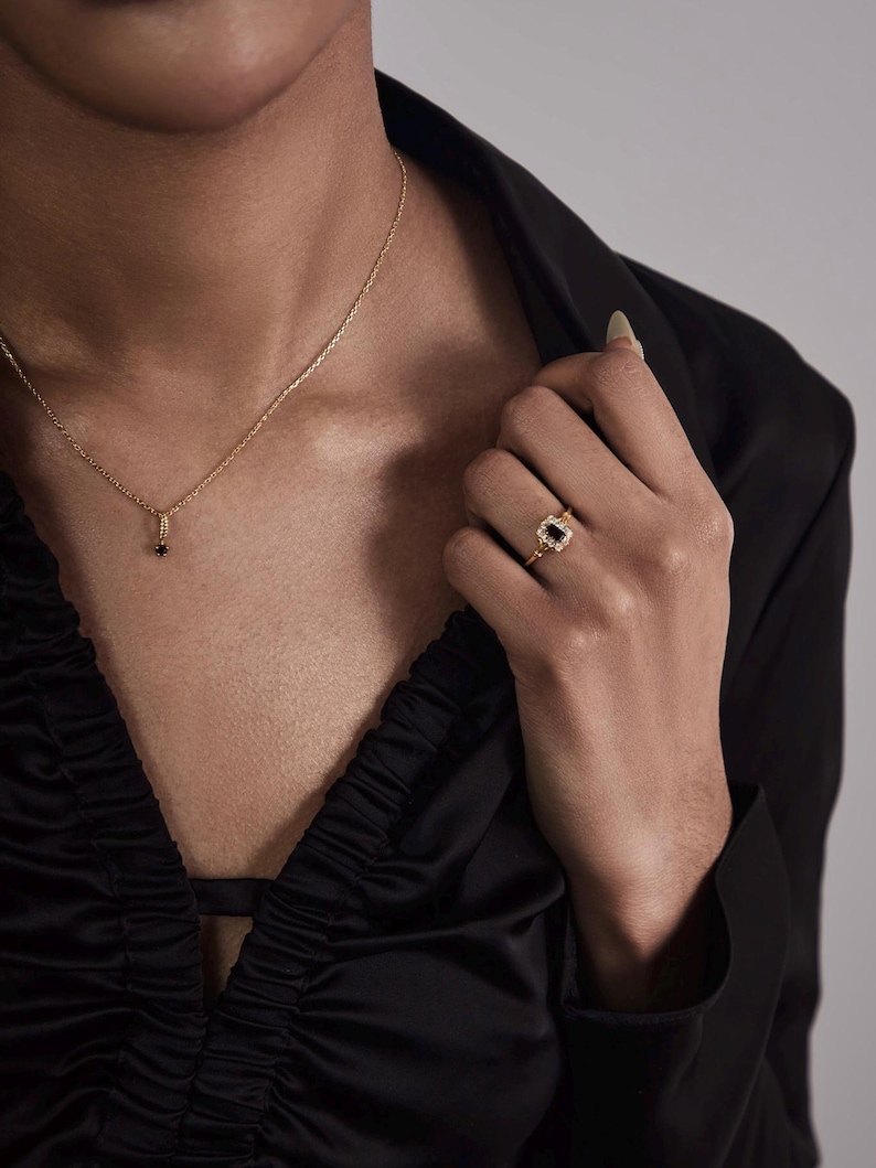 May include: A gold necklace with a small black pendant and a gold ring with a black stone. The ring has a decorative gold setting. The person is wearing a black satin top.
