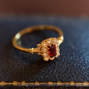 May include: A gold ring with a rectangular, deep red gemstone and small, clear stones. The ring has a thin, polished band and ornate detailing around the central stone. The ring is set against a dark background.