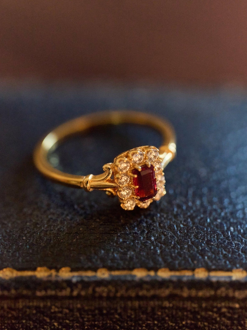 May include: A gold ring with a rectangular, deep red gemstone and small, clear stones. The ring has a thin, polished band and ornate detailing around the central stone. The ring is set against a dark background.