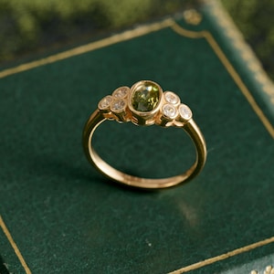 May include: A gold ring featuring a central oval-cut green gemstone, flanked by smaller, round, clear gemstones. The ring has a delicate band and is set against a dark green background.