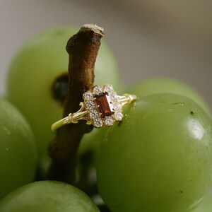 May include: A gold ring featuring a rectangular, reddish-brown gemstone, encircled by smaller, clear stones. The ring is set against a backdrop of green grapes, with the ring resting on a brown stem.