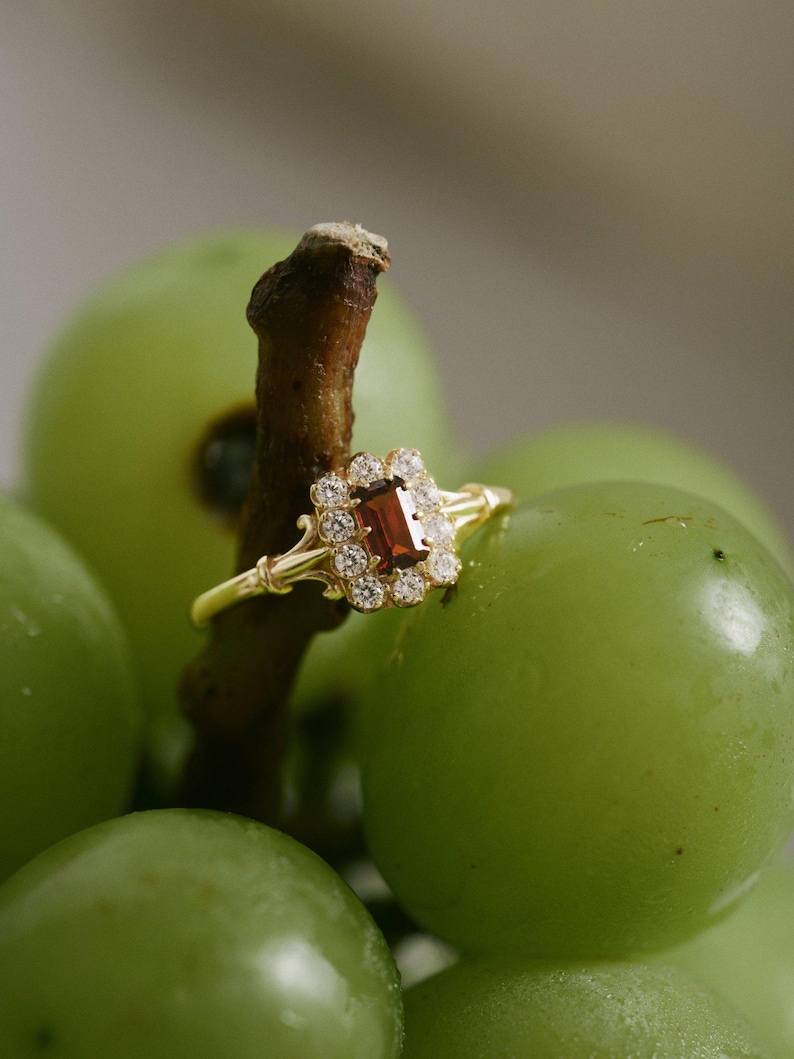 May include: A gold ring featuring a rectangular, reddish-brown gemstone, encircled by smaller, clear stones. The ring is set against a backdrop of green grapes, with the ring resting on a brown stem.