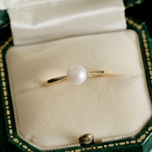 Puede incluir: Un delicado anillo de oro con una única perla blanca y redonda, presentado en un joyero de terciopelo verde con interior crema. El anillo tiene una banda delgada, que resalta la perla. El joyero es de color verde oscuro con detalles dorados.