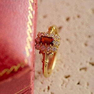 May include: A gold ring with a central rectangular, reddish-brown gemstone, encircled by smaller, clear stones. The ring has an elaborate, filigree design. The background is a soft, neutral colour, with a red box partially visible.