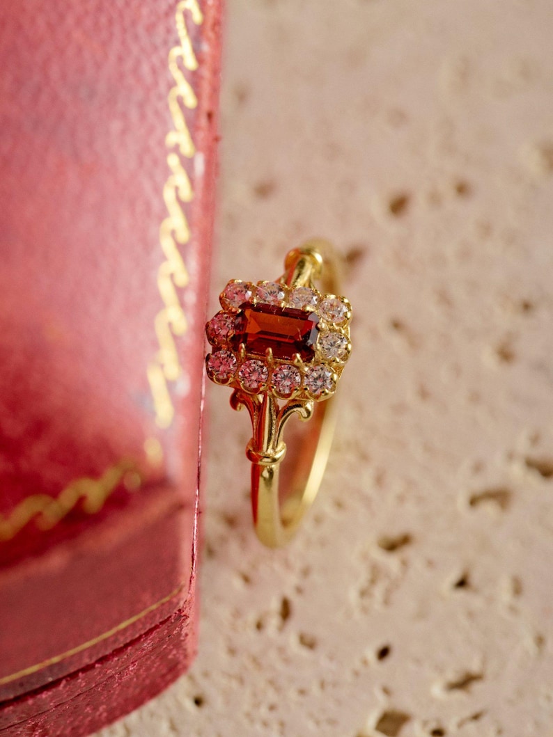 May include: A gold ring with a central rectangular, reddish-brown gemstone, encircled by smaller, clear stones. The ring has an elaborate, filigree design. The background is a soft, neutral colour, with a red box partially visible.