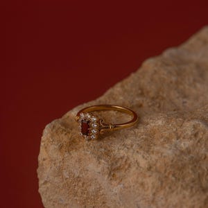 May include: A gold ring featuring a rectangular, deep red gemstone, encircled by smaller, clear stones. The ring rests on a light brown, textured rock, set against a dark red backdrop. The ring's band is slender and ornate.