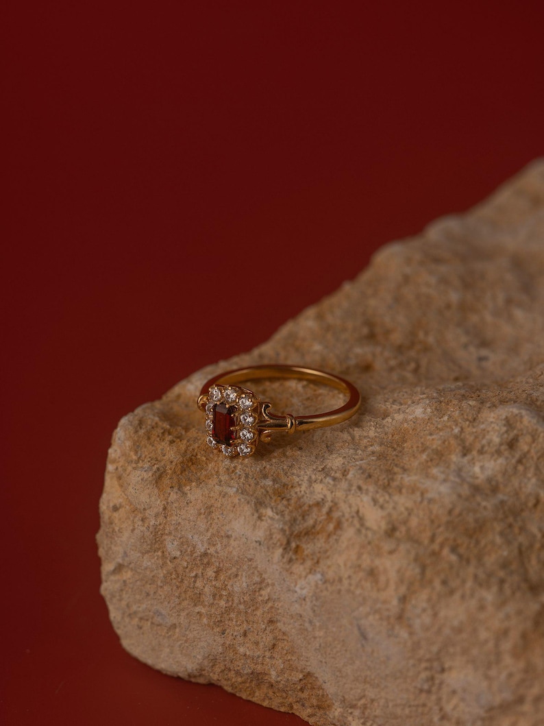 May include: A gold ring featuring a rectangular, deep red gemstone, encircled by smaller, clear stones. The ring rests on a light brown, textured rock, set against a dark red backdrop. The ring's band is slender and ornate.