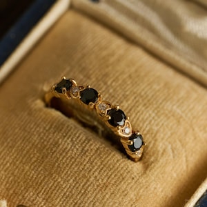 May include: A gold ring with five black gemstones and small clear stones, set in a row. The ring is displayed inside a vintage blue and gold jewelry box, showcasing the ring's design and color.
